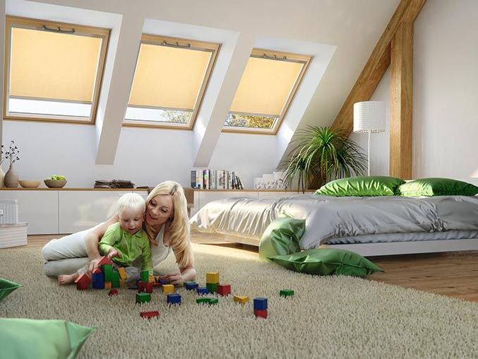 Eine Mutter spielt mit ihrem Kind auf dem Boden des Schlafzimmers, im Hintergrund 3 Dachfenster aus Holz mit fast geschlossenen Rollos innen